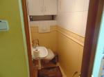 Wnętrze wąskiego prostokątnego pomieszczenia, na wprost toaleta, nad nią szafka, po lewej stronie zlewozmywak