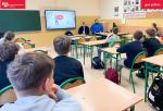 klasa szkolna, dzieci siedzą w klasie, obok tablicy wyświetlana jest prezentacja a obok niej siedzi dwóch mężczyzn